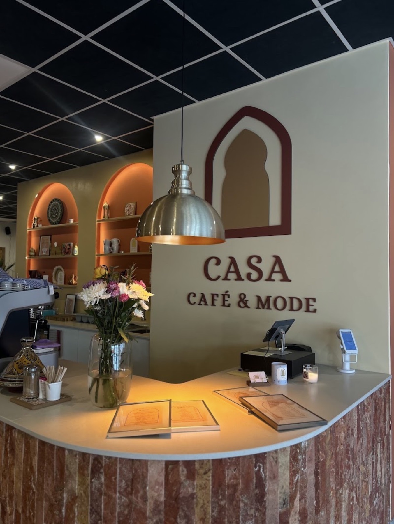 CASA Café & Mode Bar-Tresen mit beleuchteten marokkanischen Bögen, Espressomaschine und Blumenstrauß.